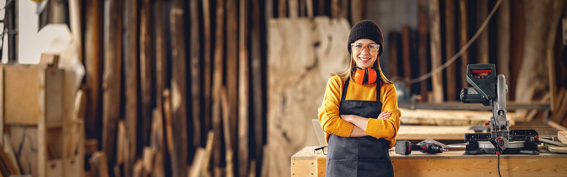 Selbstbewusst lächelnde Frau in Arbeitskleidung zur Holzverarbeitung steht in einer Tischler-Werkstatt vor einem Tisch mit Werkzeug.